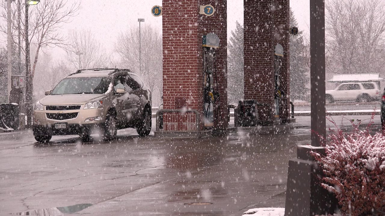 A tan crossover sits near brick gas pump columns in Westminster, Colorado as heavy snow falls over wet pavement and bare trees, with no visible people.