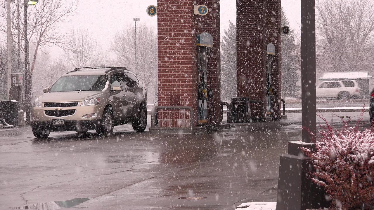 The tan crossover remains by the pump island while dense snow streaks across the Westminster gas station forecourt and a pale vehicle passes in the background, with no visible people.