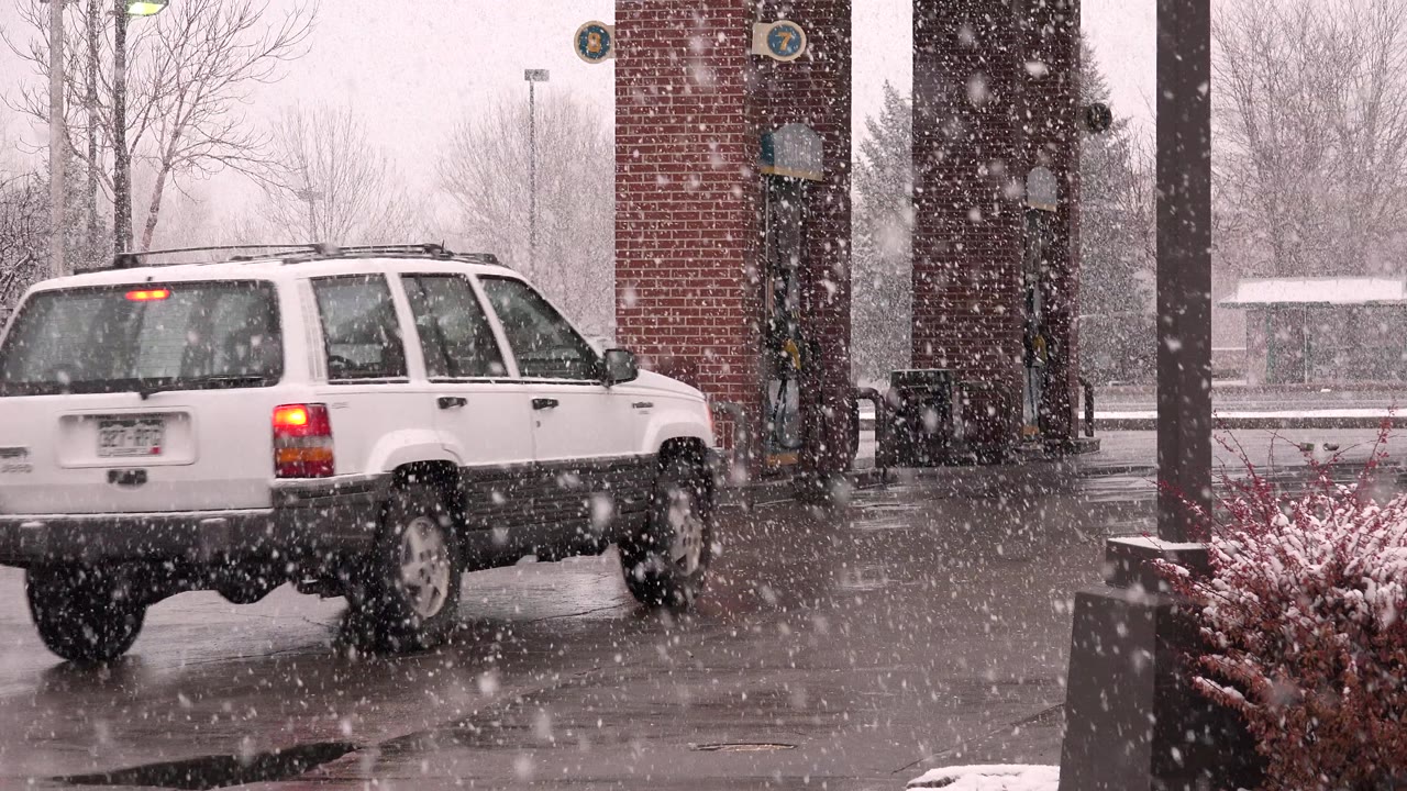 A white SUV moves left to right through falling snow in Westminster, Colorado, with wet pavement, brick columns, bare trees, and no clearly visible people.