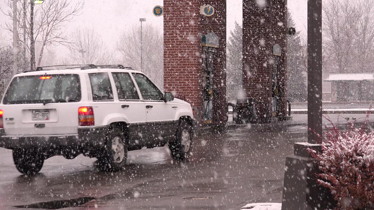 The white SUV continues across the frame in Westminster, Colorado, passing the brick pump area as snow thickens against a gray winter background.