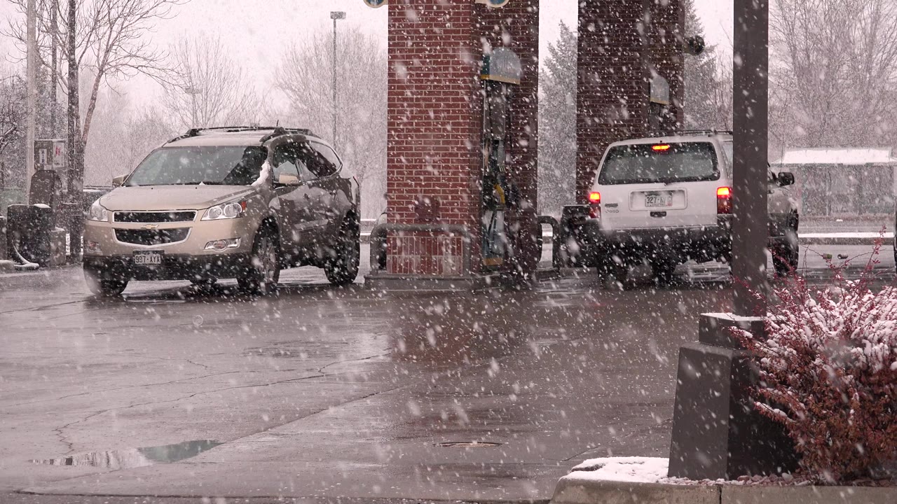 Opening frame of a Westminster, Colorado gas station during heavy snowfall, with a beige crossover at left, a white minivan near brick pump columns, wet pavement, bare trees, and no visible people.