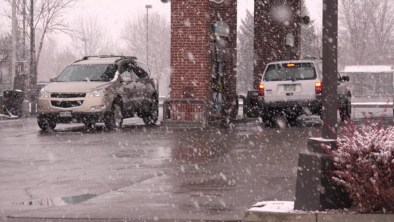 A white minivan sits near the gas station pumps in Westminster, Colorado, with falling snow crossing brick columns, a wet apron, bare winter trees, and no visible people.