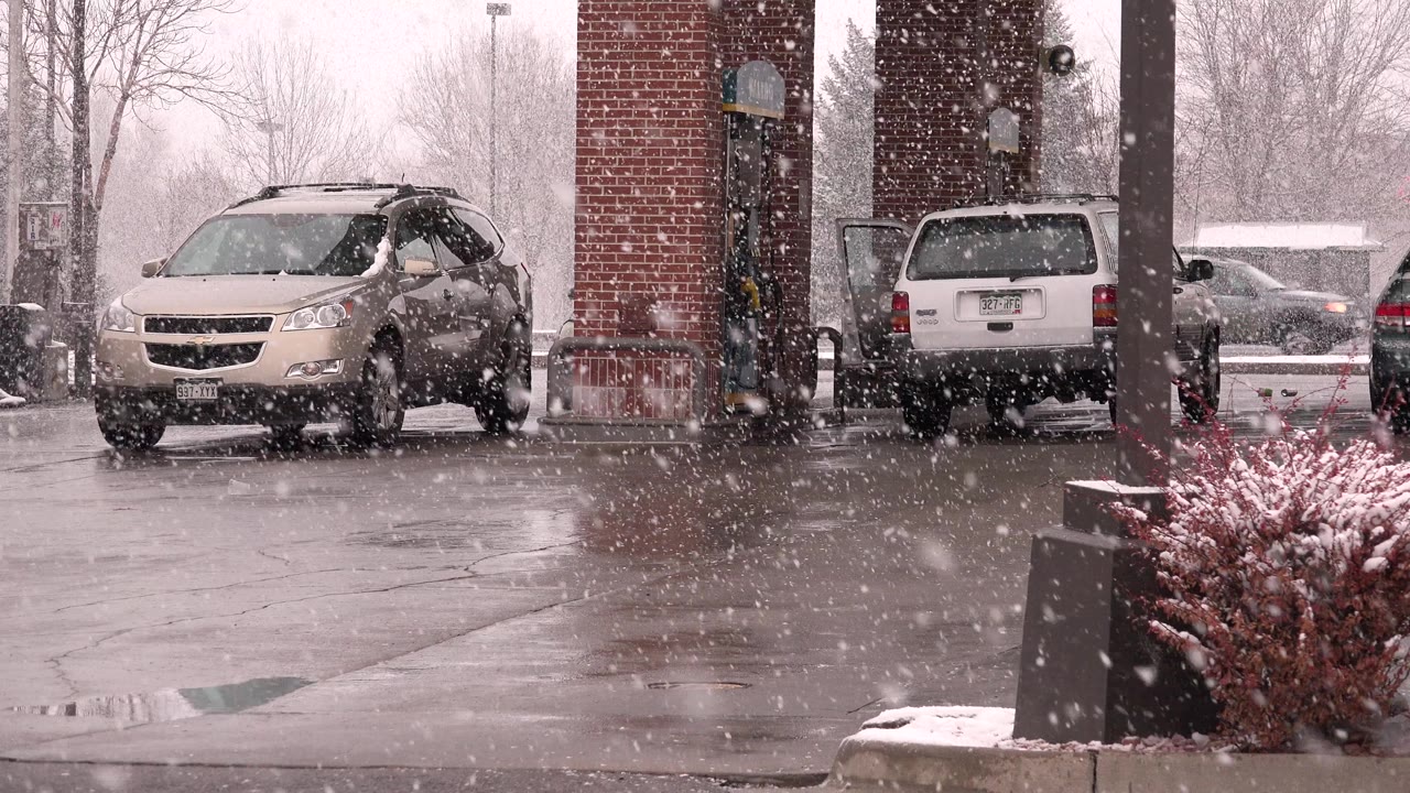 Heavy snow falling over parked vehicles beside a brick building.