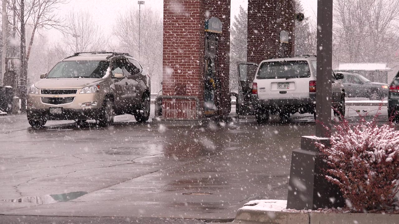 A Westminster, Colorado parking area is seen through heavy snow, with a beige minivan at left, a white SUV near the brick column, and wet pavement reflecting the overcast light.