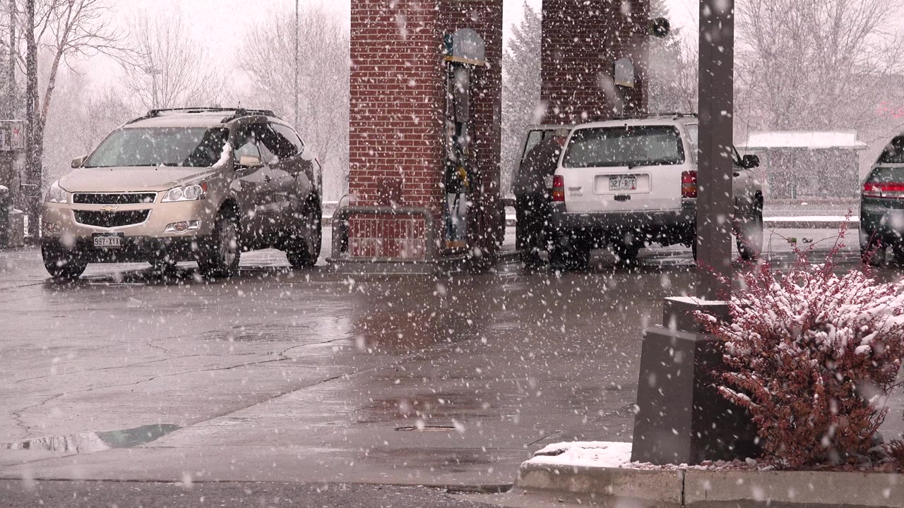 Heavy snow falls at a Westminster, Colorado gas station with a tan crossover on the left, a white SUV by the brick pump island, wet pavement, and no visible people.