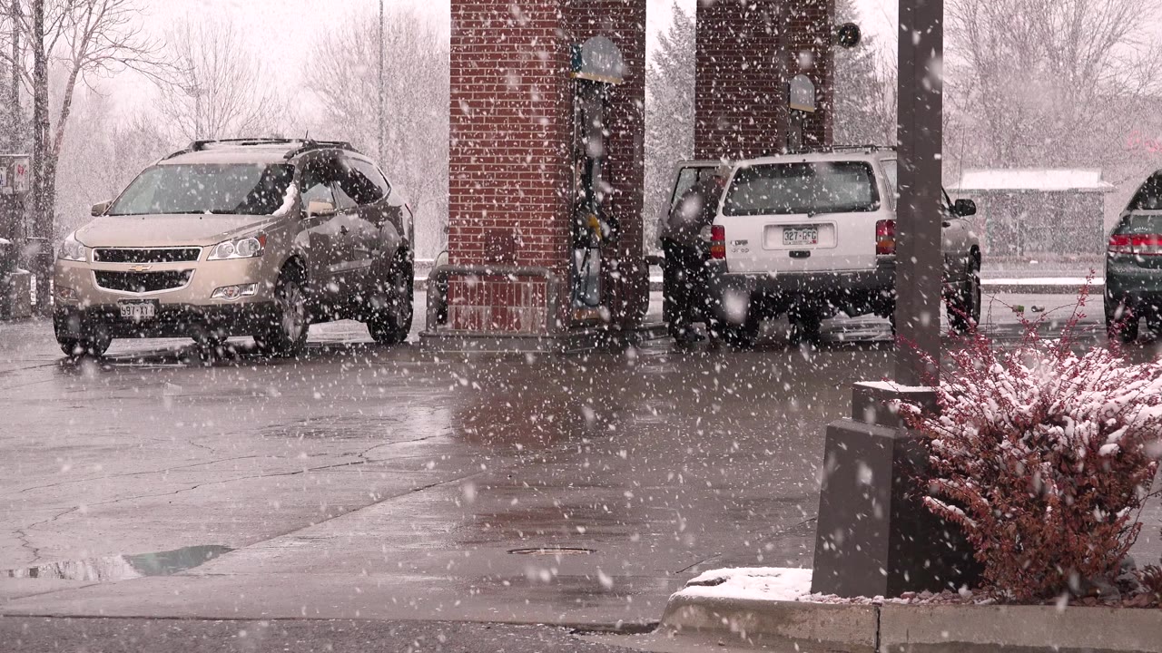 Snow continues across the Westminster, Colorado forecourt as the tan crossover remains left of the brick pump island and the white SUV sits near the pumps, with no visible people.