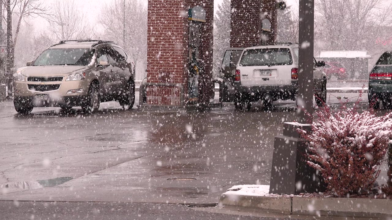 At 0:01 in Westminster, Colorado, larger foreground flakes cross the view while the tan SUV, white SUV, brick column, wet pavement, and planter remain in frame, with no visible people.