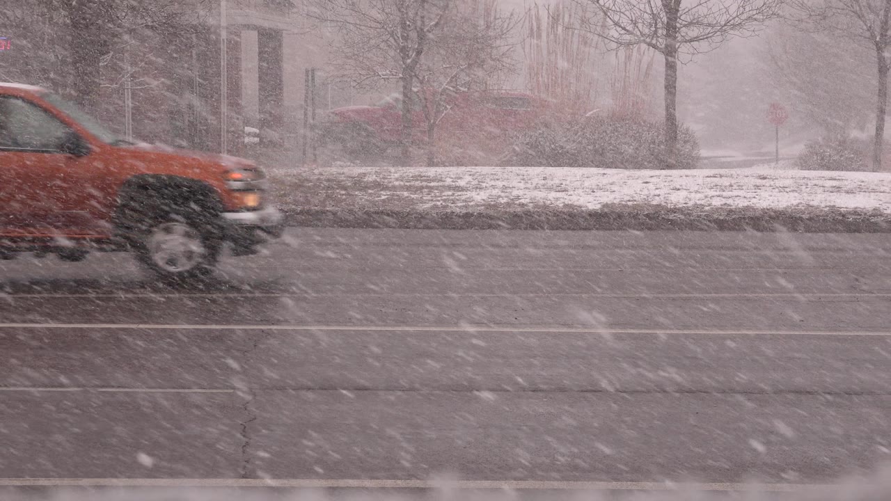 An orange pickup truck moves across a snow-swept roadway in Westminster, Colorado, with bare trees and muted buildings behind it and no visible people.