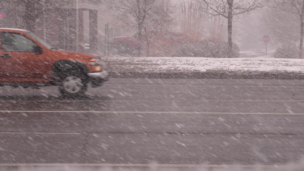 The orange pickup is centered farther down the Westminster roadway as dense wind-driven snow streaks across the lanes and no people are visible.