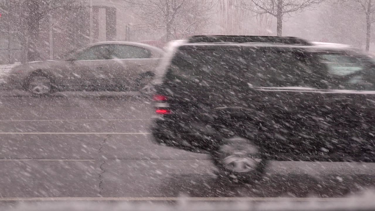 A dark SUV crosses the right side of the Westminster, Colorado road during heavy snowfall, with a light car partly visible behind it and no visible people.