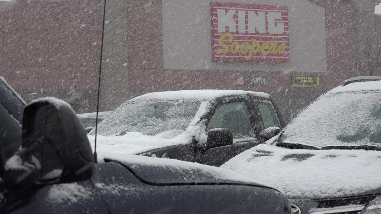 Heavy snow blows across parked vehicles in a storefront parking area.