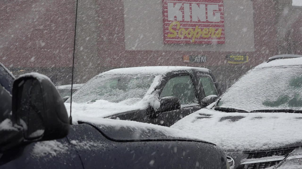 Snow falling over parked cars outside a King Soopers storefront.