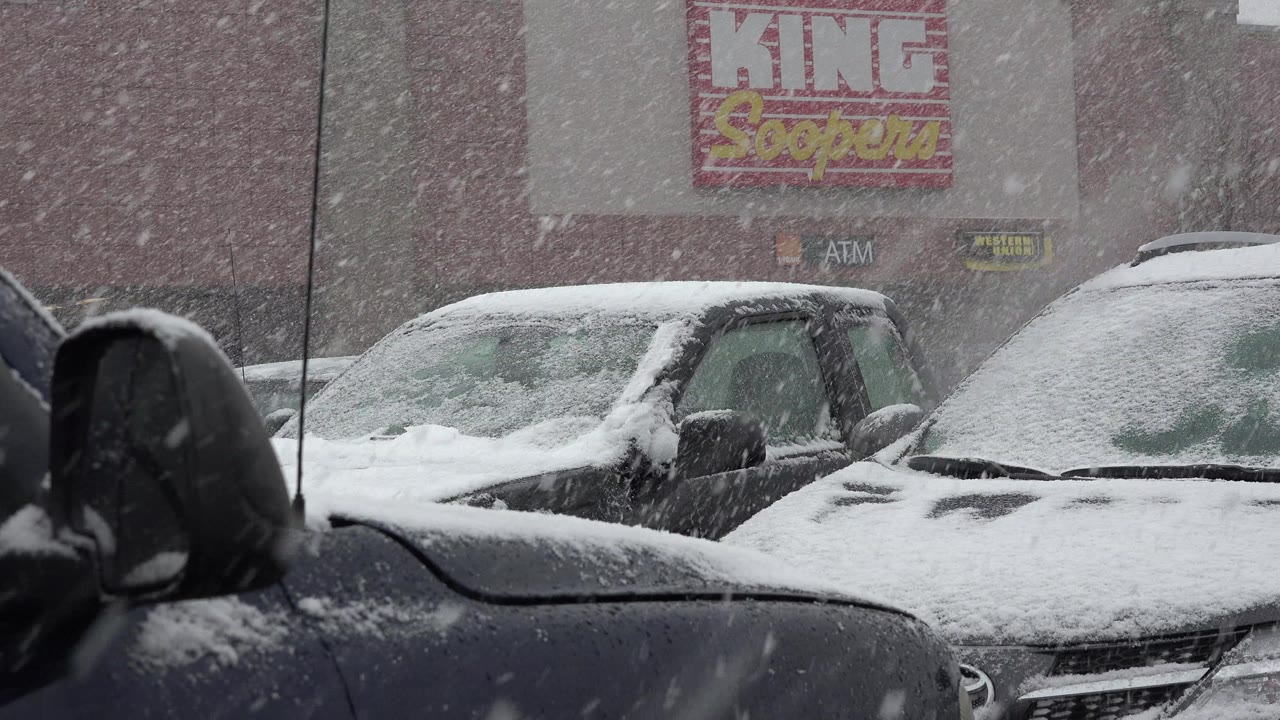 A tight Westminster, Colorado parking-lot frame shows heavy snowflakes crossing the view, two snow-capped cars in the midground, storefront signage above, and no visible people.