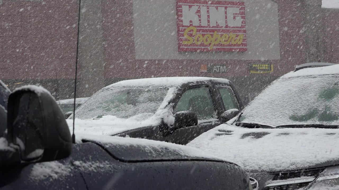 At 0:00 in Westminster, Colorado, heavy snow falls across parked cars and trucks in a retail lot, with King Soopers signage in the background and no visible people.