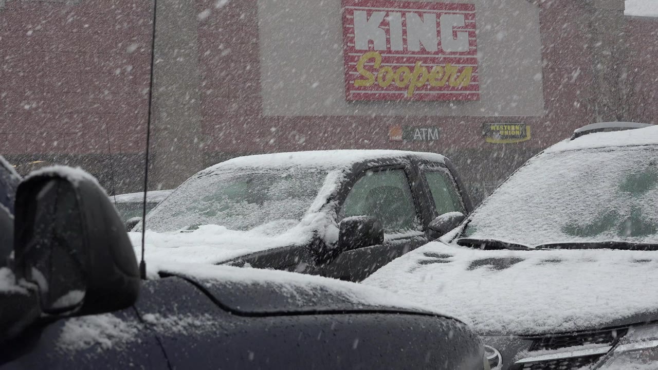 At 0:01, snow streaks across a close view of parked vehicles in Westminster, Colorado, with white buildup on windshields and hoods and no visible people.