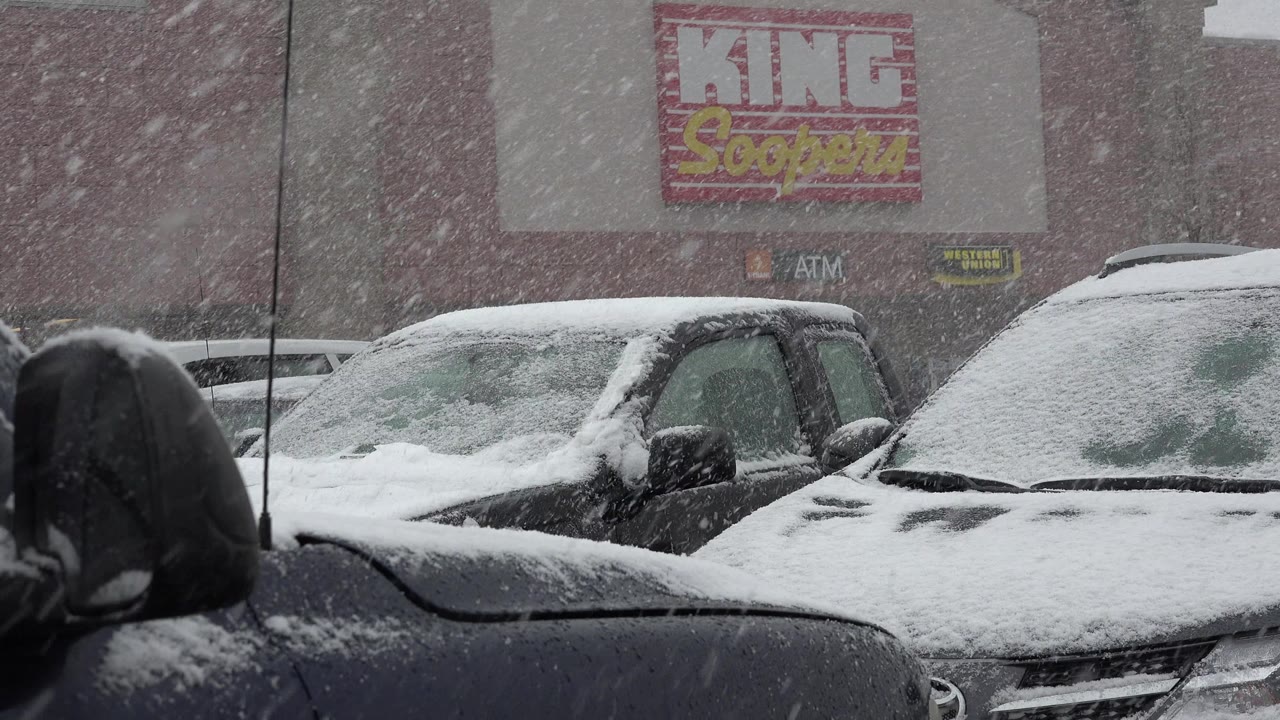 At 0:01, the snowy parking lot remains framed from behind a dark foreground vehicle, with coated windshields, a store wall, King Soopers signage, and no visible people.