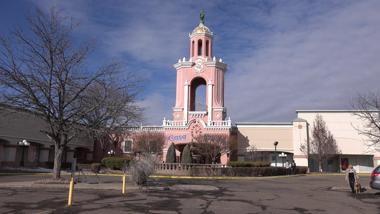 Wide ground-level view in Denver, Colorado of a pink and cream building with a tall arched tower, bare trees, bollards, hedges, and no clearly identifiable people in the first frame.