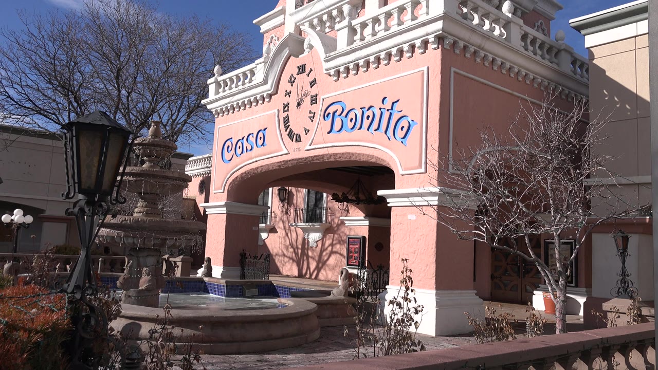 Denver frame with the Casa Bonita sign centered above an arched entrance, a tiered fountain at left, bare branches at right, and small distant people near the doorway.