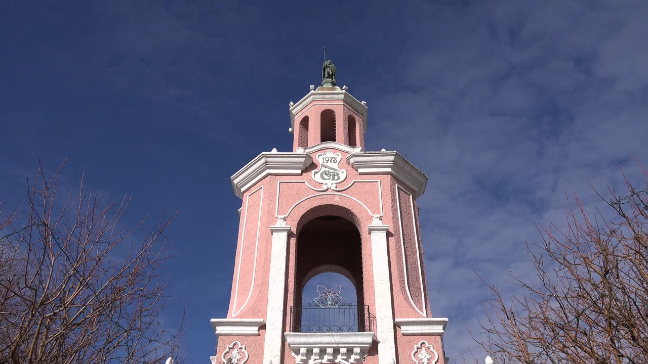 Denver frame showing the upper section of a pink clock tower with white trim, an arched opening, blue sky, bare tree branches, and no visible people.