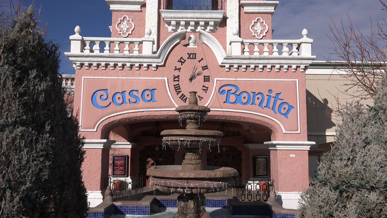 Denver, Colorado exterior view of the pink and white Casa Bonita facade with blue lettering, a front fountain, trees at both sides, and no visible people.