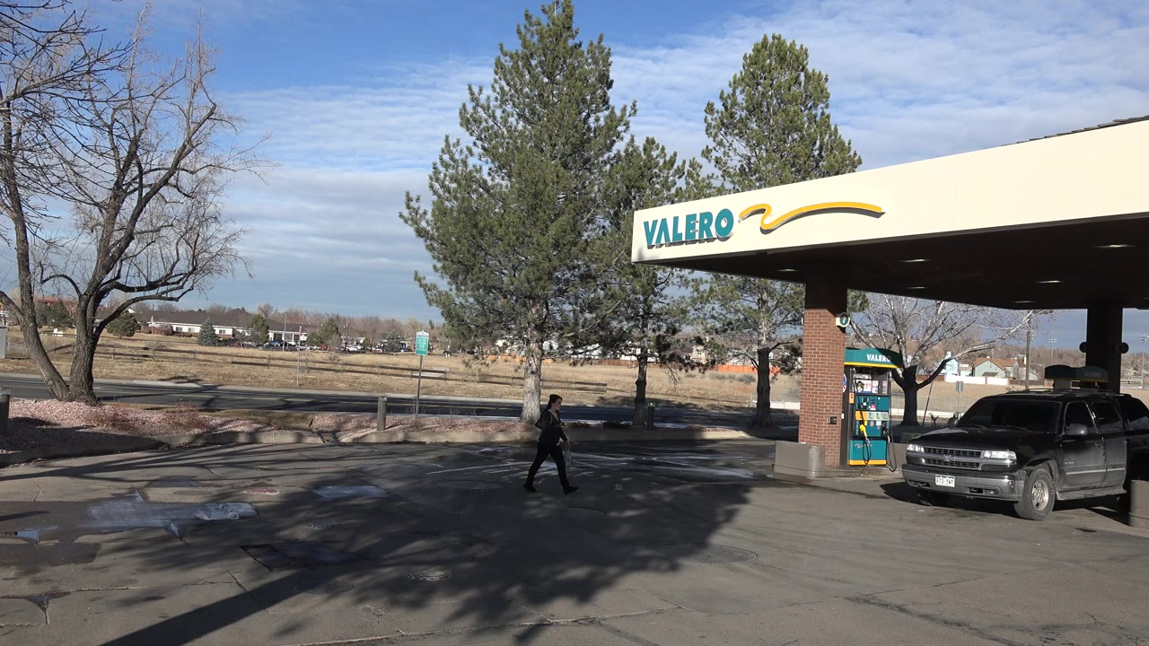 Denver, Colorado gas station forecourt with a Valero canopy, dark SUV at the pump, leafless trees, open roadside background, and one distant person walking across the pavement.