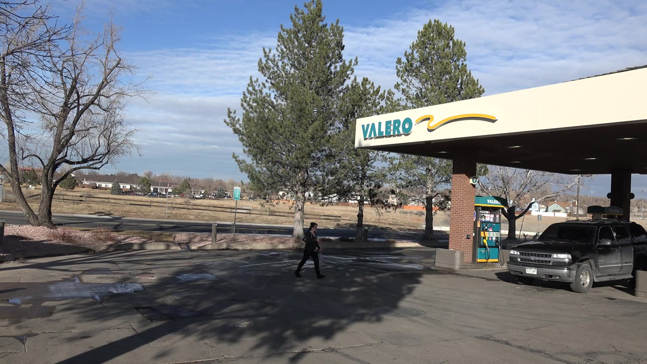 Denver gas station scene with the Valero sign high on the canopy, a dark SUV under the pump island, long tree shadows on the pavement, and a small walking figure near the left side.