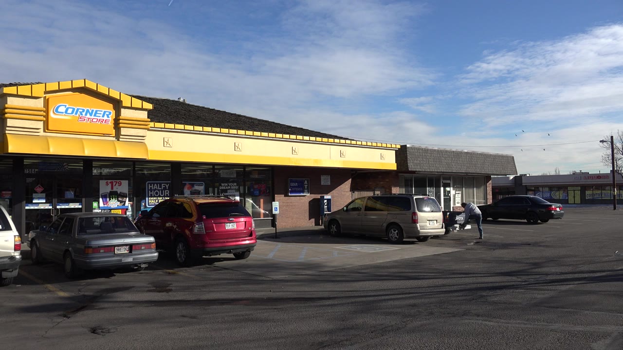 Wide Denver, Colorado frame of a yellow Corner Store facade, parked cars, glass storefront windows, asphalt foreground, blue sky, and a small person near a vehicle at the right.