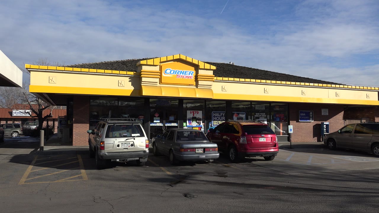 Convenience store storefront beside parked cars and a gas station canopy.