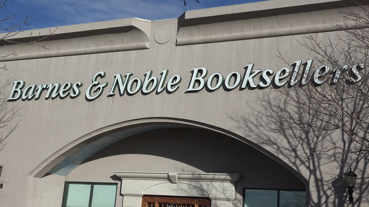 Close storefront view in Denver showing the Barnes & Noble Booksellers lettering centered over the arch, with tree shadows crossing the pale facade and no visible people.