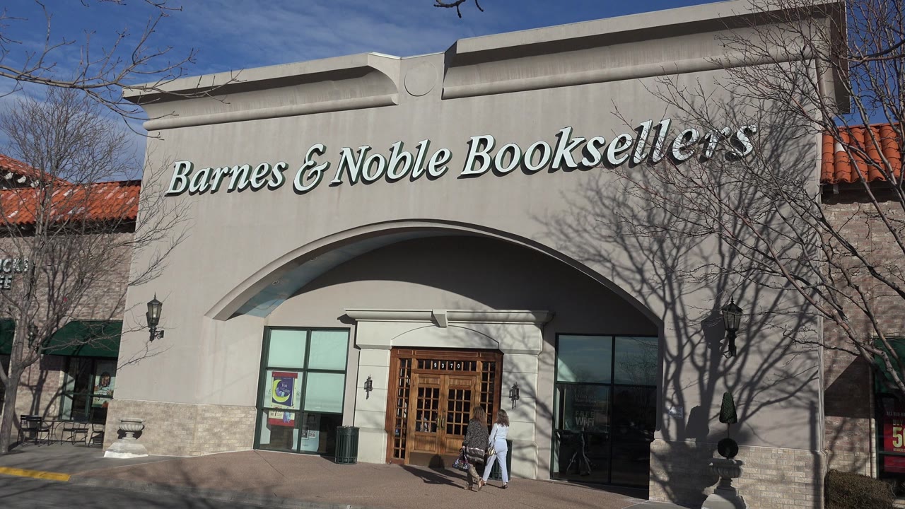 Denver, Colorado frame showing the Barnes & Noble Booksellers sign above an arched storefront, with two people walking near the wooden entrance doors.