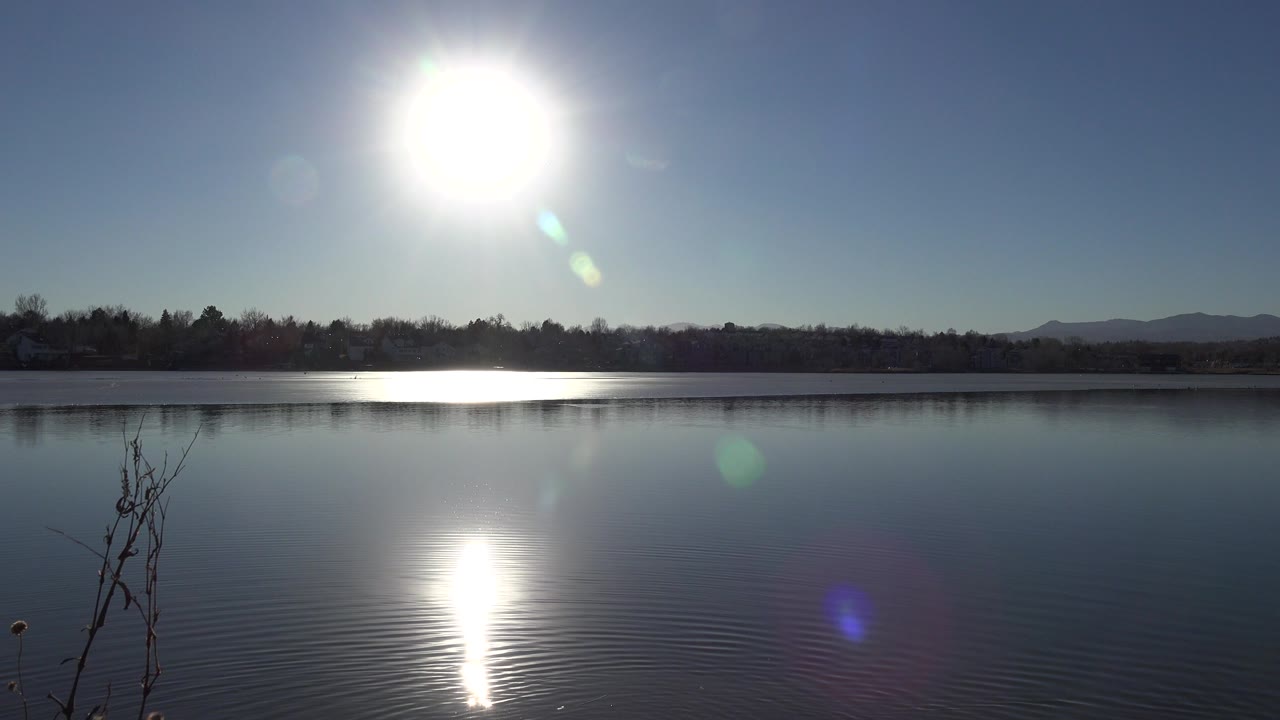 At 0:00 in Westminster, Colorado, the sun hangs high over a calm lake, reflecting as a white vertical streak on the water with a dark tree line and low hills beyond, and no visible people.