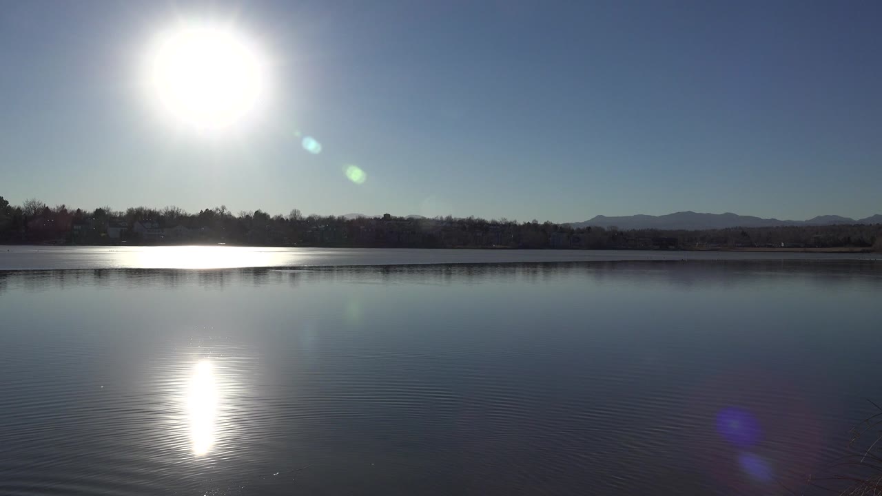At 0:01, the sun sits high above the Westminster lake while green lens flare spots angle downward over the water and the far tree line remains dark against low hills, with no visible people.