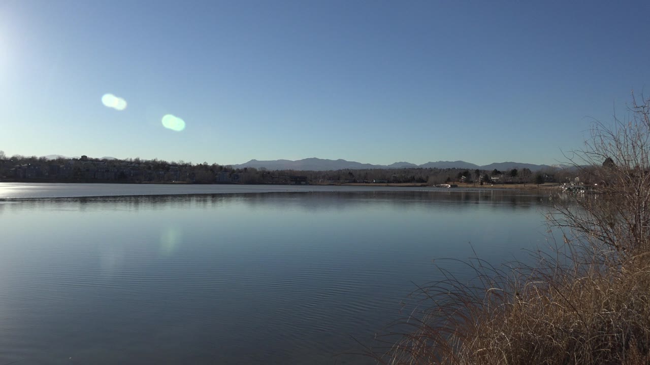 Blue lake with brown shoreline brush and distant hills under clear sky.