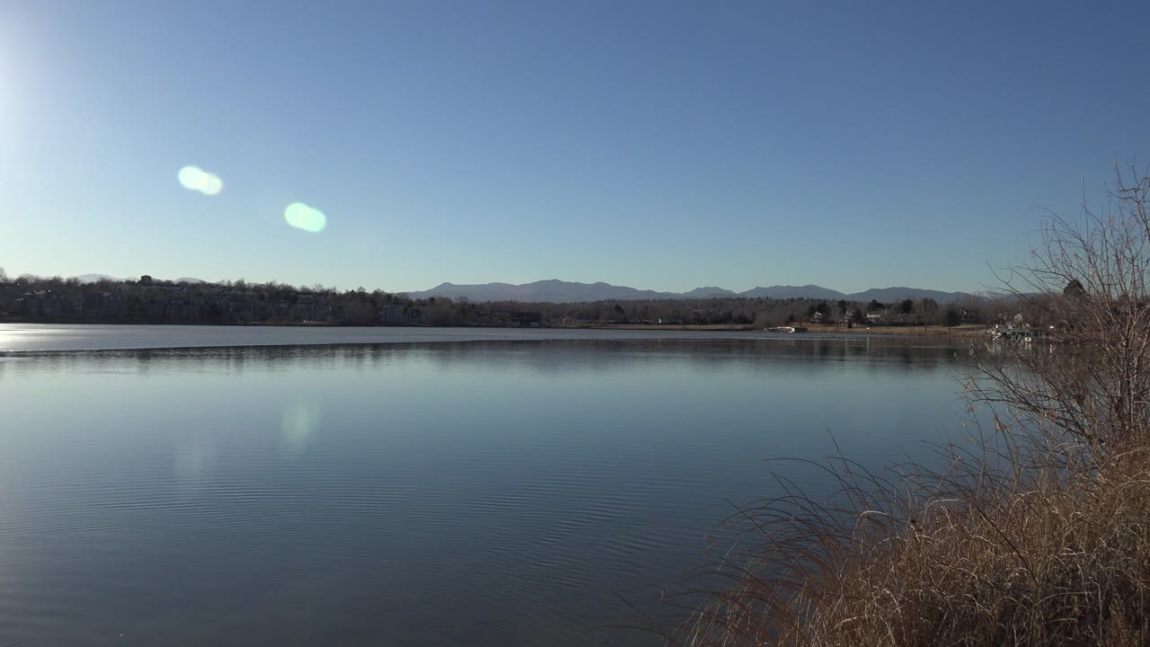 Westminster, Colorado lake frame with calm blue water across the center, brown brush at the right edge, low hills on the far horizon, sky glare, and no visible people.