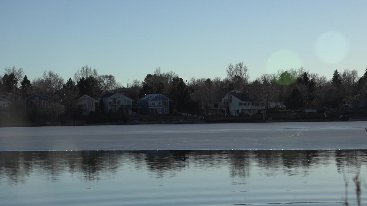 Second frame holds on the Westminster lake with broad reflective water, silhouetted houses and trees on the far bank, and a bright circular lens flare in the sky, with no clearly visible people.