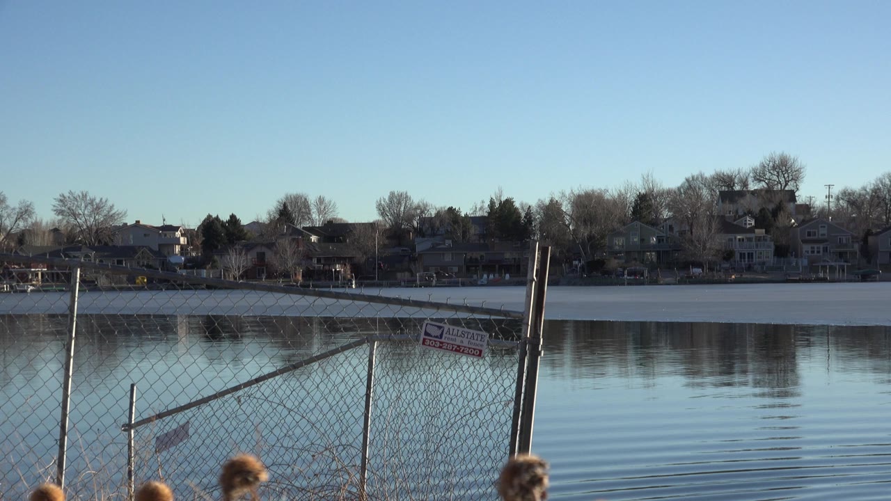 Westminster, Colorado lake view with the fence angled across the foreground, soft ripples on the water, distant houses in shadow, and no visible people.