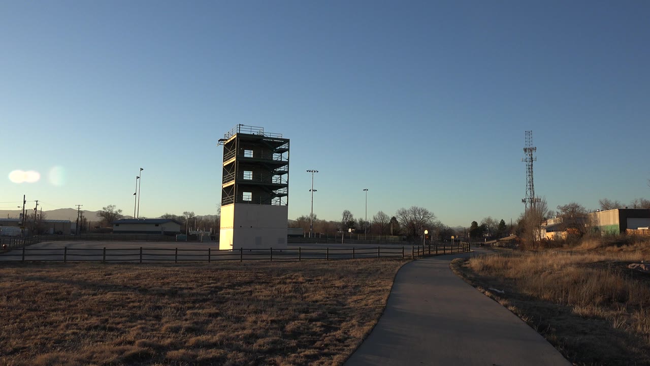 Opening frame of a Westminster, Colorado construction site with a half-finished tower left of center, a paved path in the foreground, dry grass, fencing, distant hills, and no visible people.