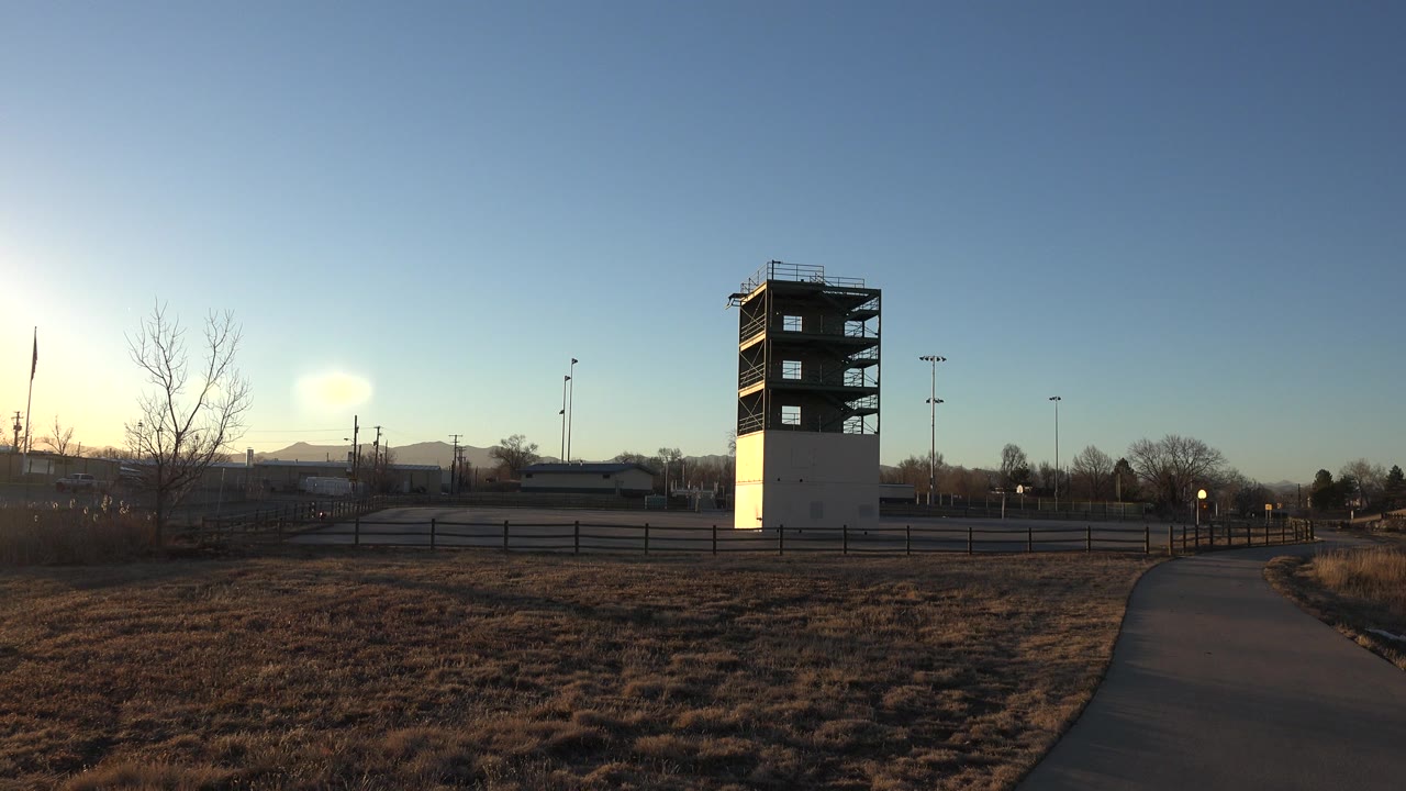 The Westminster tower remains centered beyond the paved path, with dry grass in the foreground, a fence line behind it, and no visible people.