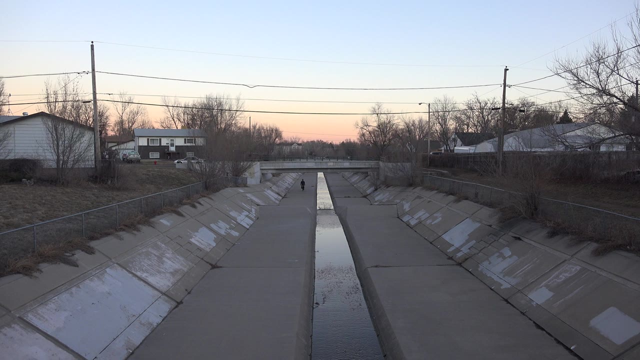 Concrete drainage ditch with narrow water channel, houses, trees, and a distant walker.