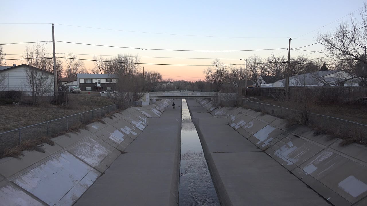 The Westminster drainage canal remains centered, with reflective water running between grey concrete slopes, houses beyond the channel, bare trees, and a very small distant walker.