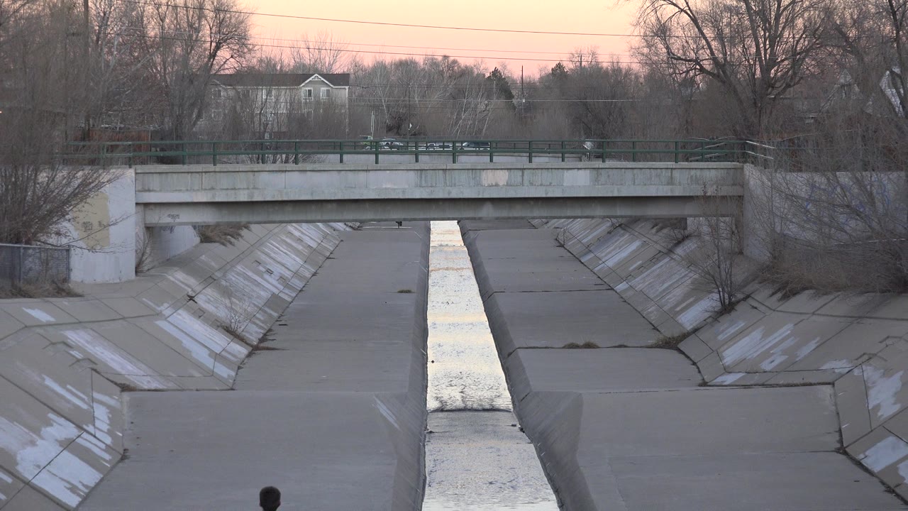 Concrete drainage channel with narrow flowing water and bridge in background.