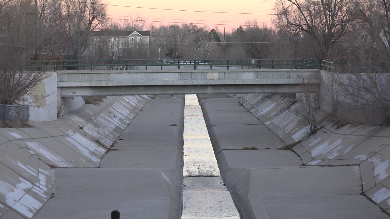 Westminster, Colorado waterway centered in a sloped concrete channel below a bridge, with winter brush, muted evening light, and a small partial view of a jogger near the bottom edge.