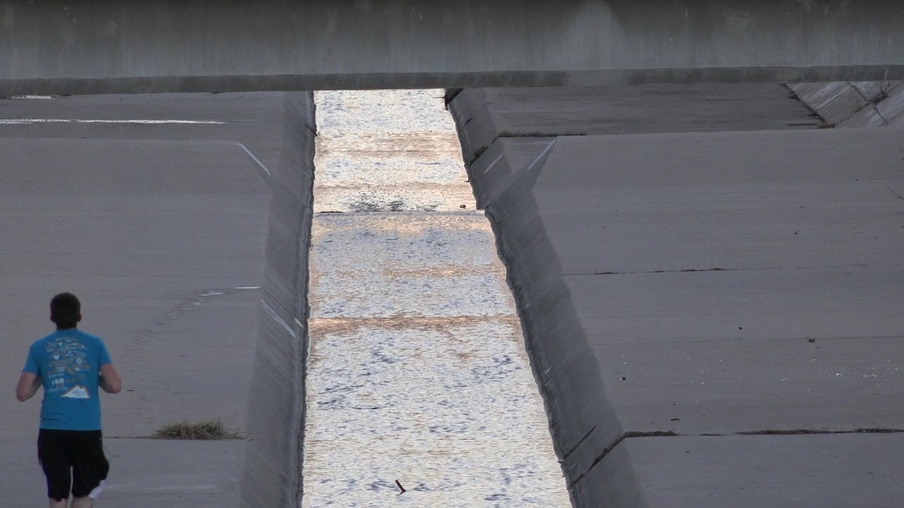 One second in, the runner remains left of center while the bright water strip and sloped concrete bank fill the middle of the Westminster channel view.