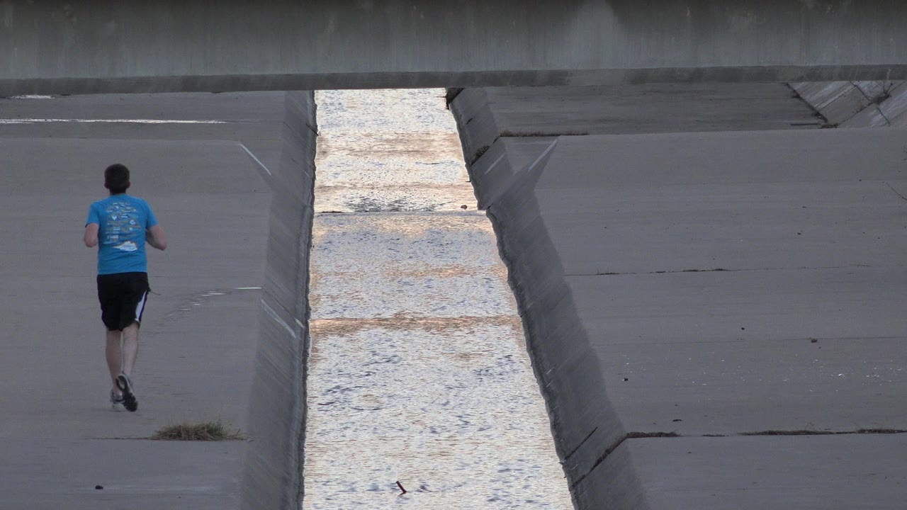 Man jogging away beside a concrete water channel under a bridge.