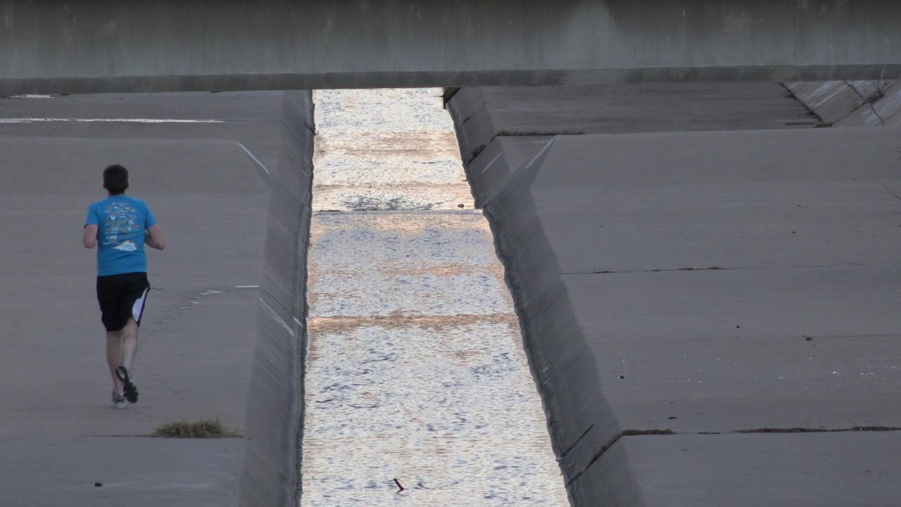 One second in, the runner is farther from camera on the Westminster concrete channel path, with the pale water strip cutting through the frame beside him.