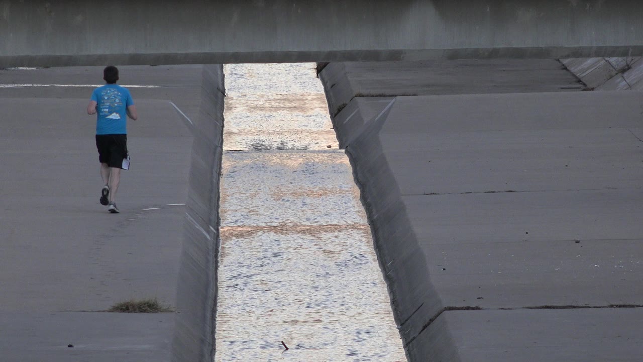 At 0:00, a man in a blue shirt jogs away beside a pale water channel in Westminster, Colorado, with a low bridge crossing the top of the frame.