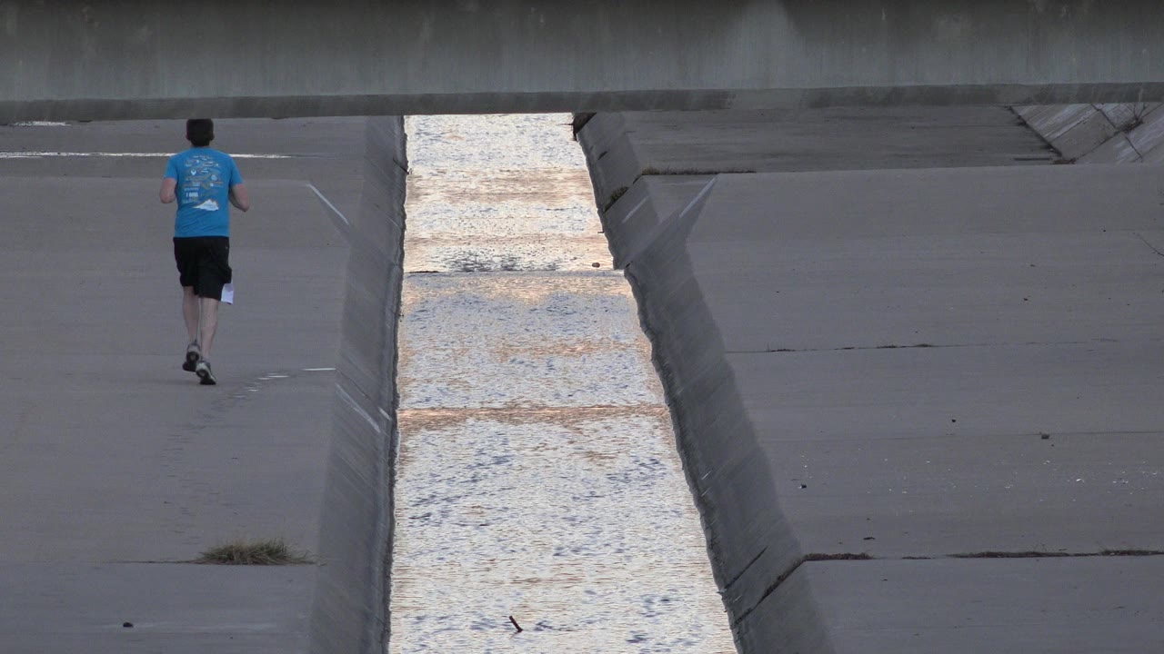 At 0:01, the runner has moved farther along the concrete path beside the reflective water strip in Westminster, Colorado.