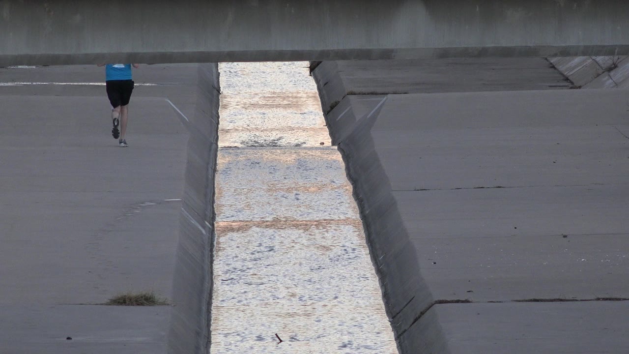 A distant runner appears near the top of a gray concrete canal scene in Westminster, Colorado, with reflective water running vertically through the frame.