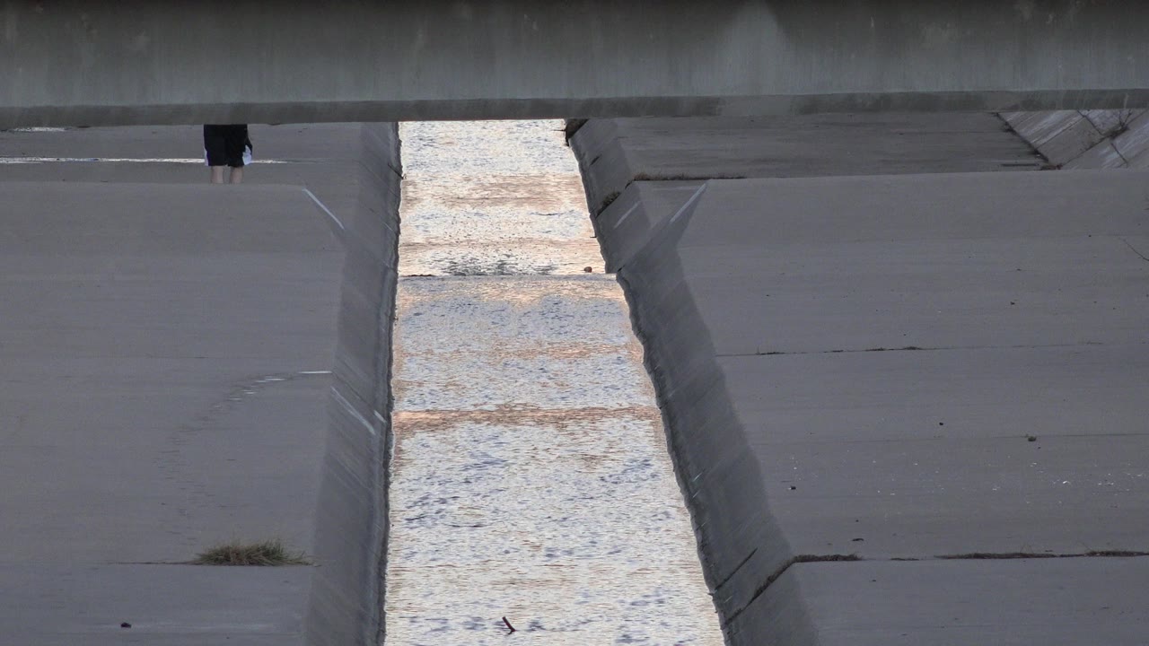 A narrow reflective water channel runs between gray concrete banks in Westminster, Colorado, with a partially visible walker cropped at the top edge.
