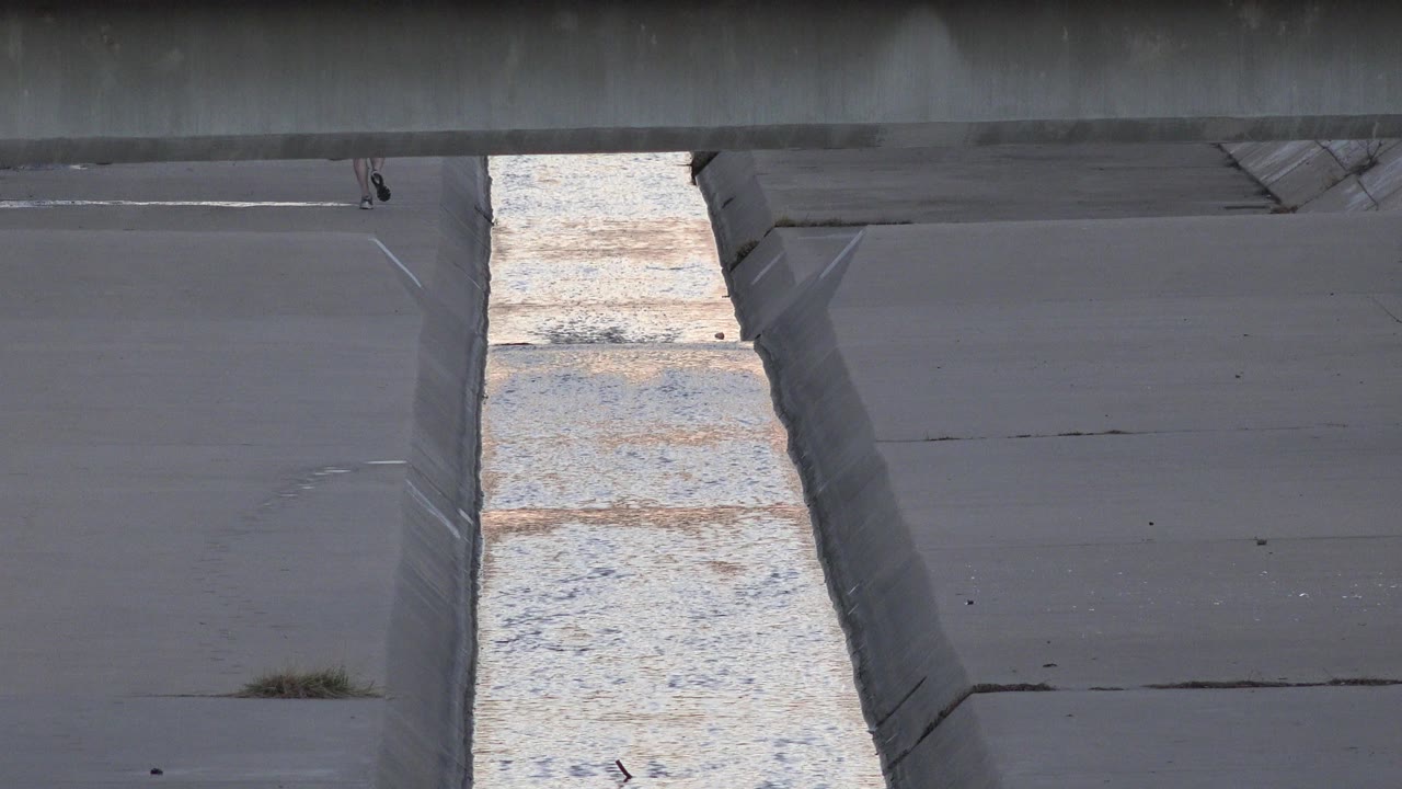 Concrete channel beneath a dark bridge beam with rippling water below.
